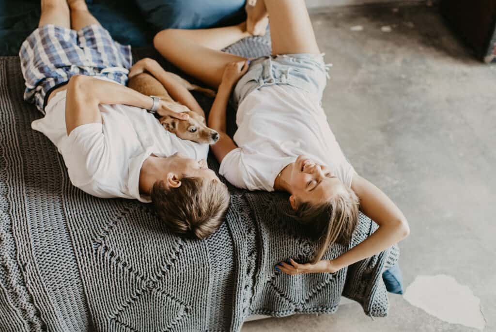 A couple lays on their backs in a grey bed with a dog between them