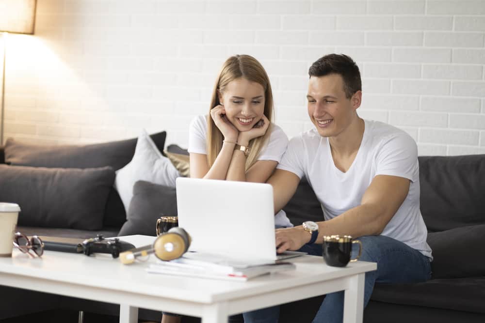A couple sits on the couch smiling while on the computer.