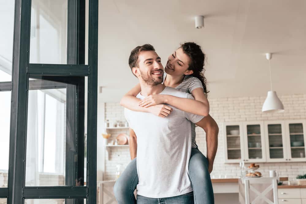 A man holds a woman on his back as they laugh in the kitchen. 