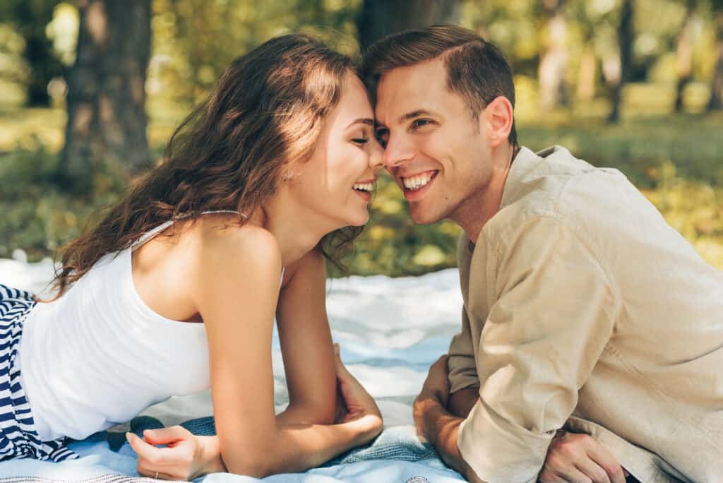 A man and woman are nose to nose while sitting outdoors on a blanket.