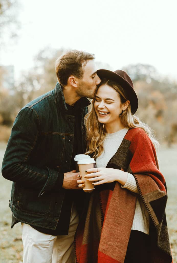 A man kisses a woman\'s head outdoors. They both hold coffee cups.