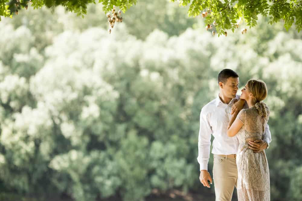 A couple embraces one another in a forest. The woman wears a lace dress.