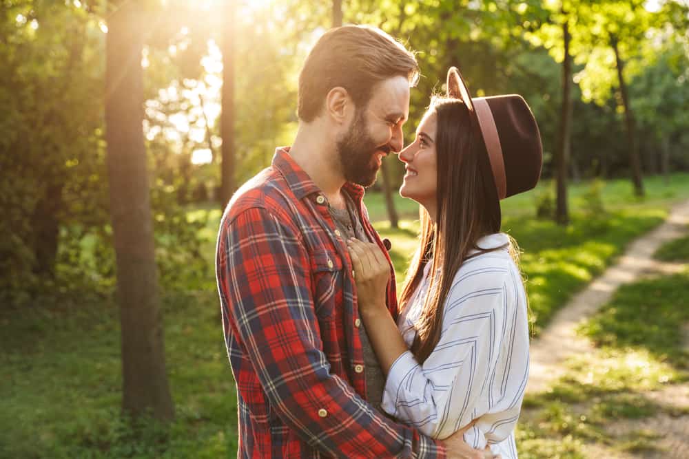 A cheerful couple is nose to nose about to kiss in a forest.