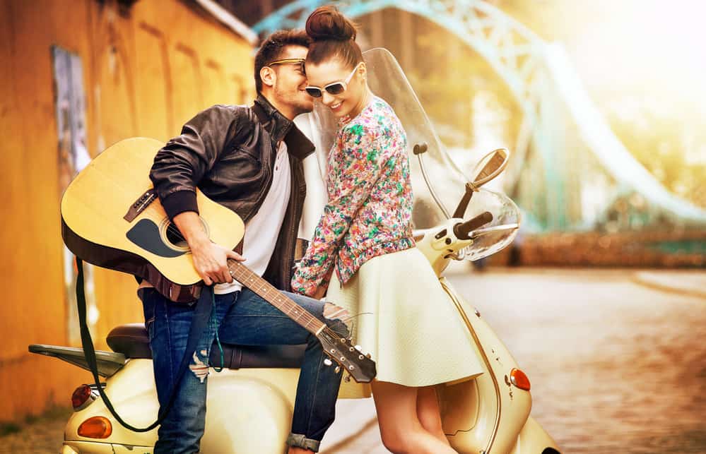 A man kisses a woman's cheek while sitting on a vespa. He's holding a guitar and she is wearing white sunglasses.
