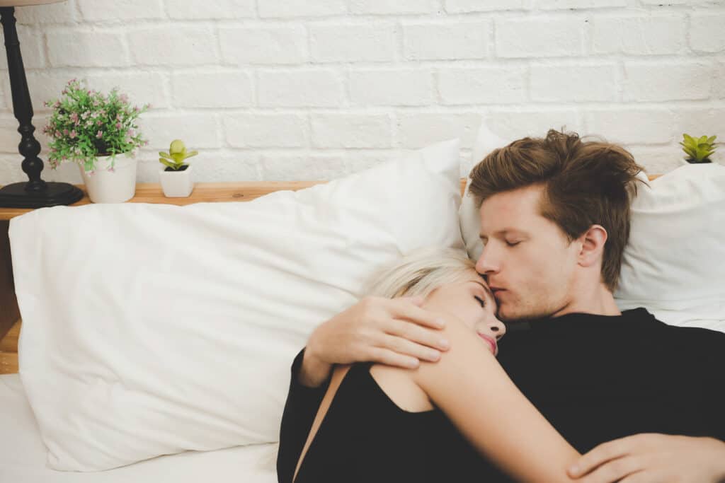 A man kisses his girlfriend\'s forehead while she cuddles him in a white bed. They both wear black shirts.