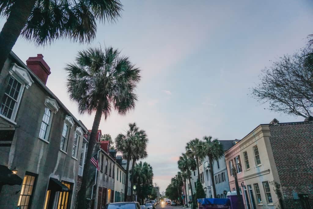 Cars line a street with pastel-colored homes and palm trees as the sun sets.