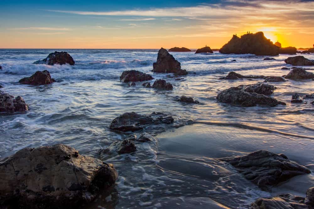 Waves crash in the water with rocks under a blue and orange sky.