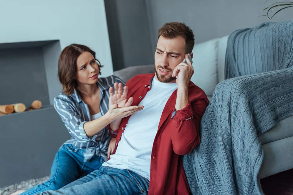 A couple is arguing on a couch. He is on his phone pushing her away as she gestures towards him.