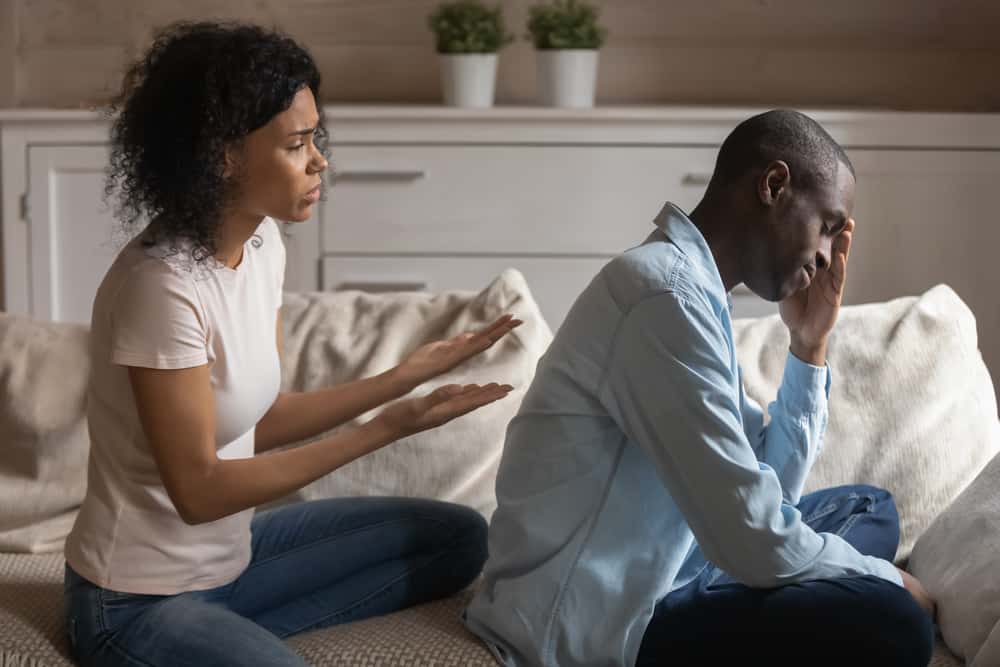 black woman and man in argumentative pose, she is gesturing behind his back towards him while he has his head in one hand