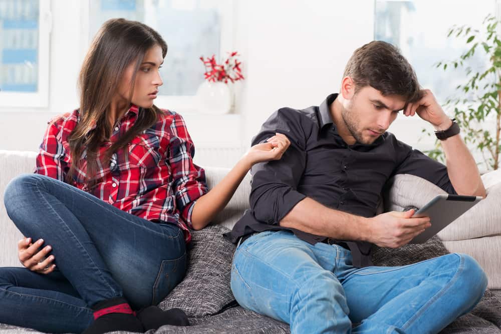 A woman looks at a man on a couch while he stares at his phone.