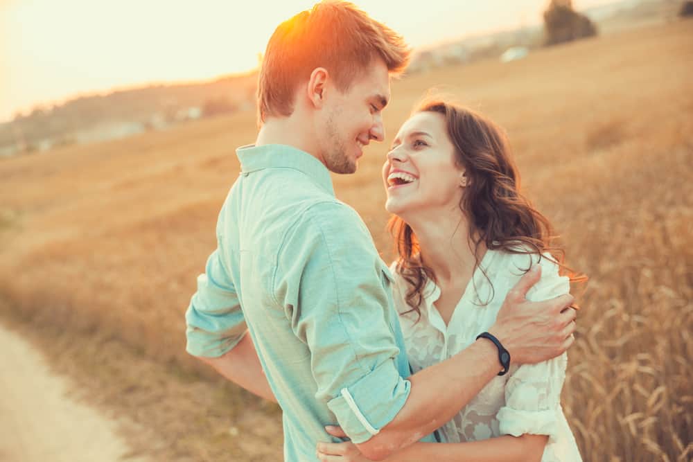 A couple is embracing one another in a field as the sun sets behind them.