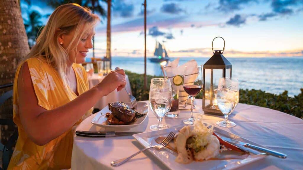A woman dives into a piece of steak at a candlelit dinner by the ocean.