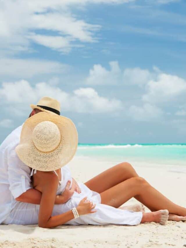romantic beach getaways in usa - couple sitting on white sandy beach near turquoise water, they are laying together and looking away from the camera. both wear sun hats