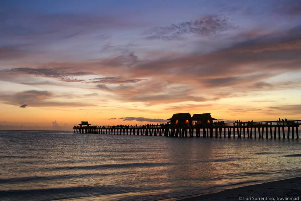 A pier stretches across the ocean under a purple/orange sky as the sun sets.