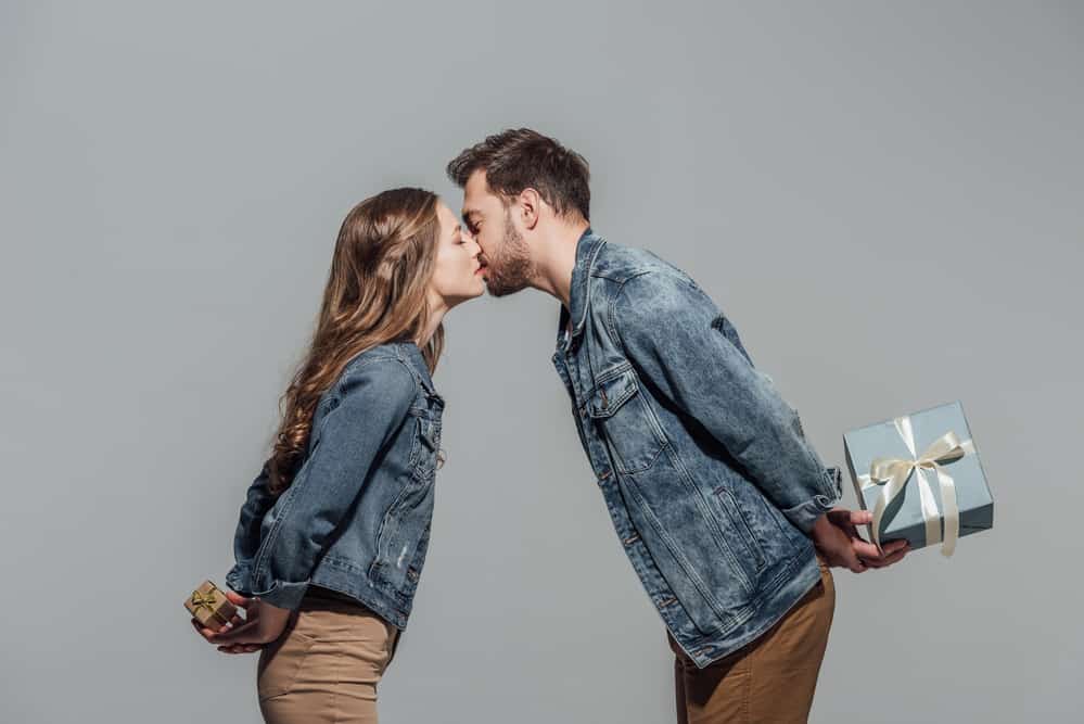 Image of a couple in front of a grey background. They are leaning in to kiss on the lips, holding gifts behind their back. Both are wearing denim jackets.