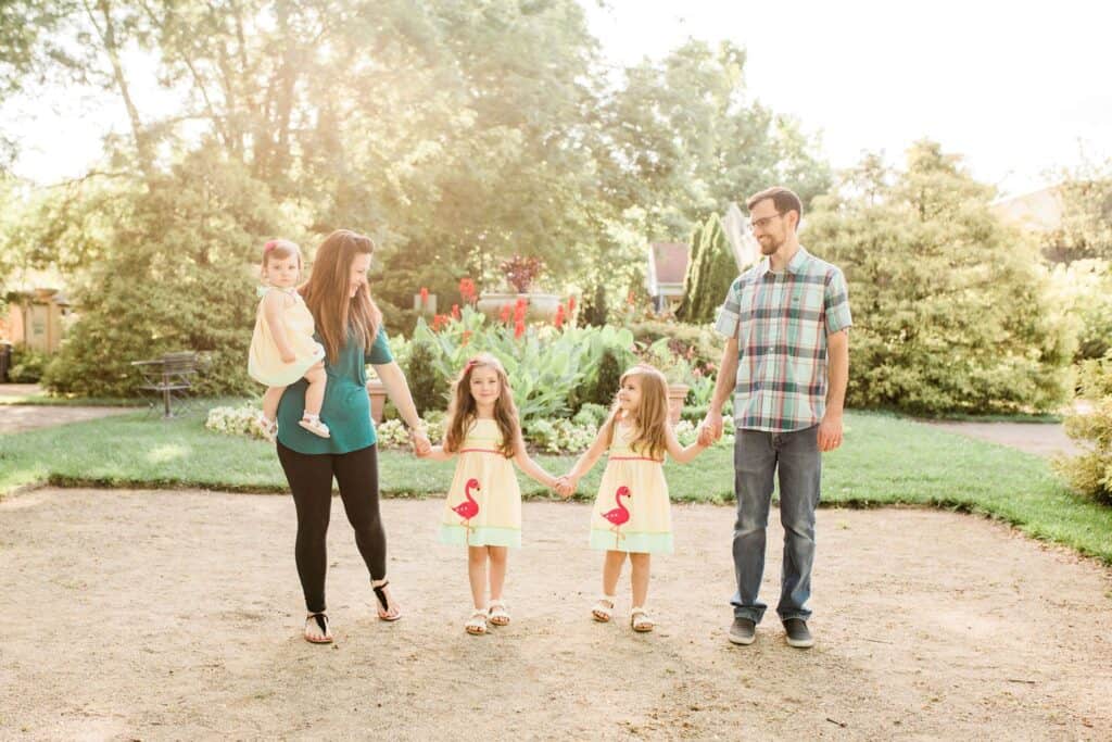 A couple stands while holding their daughters\' hands in a garden.