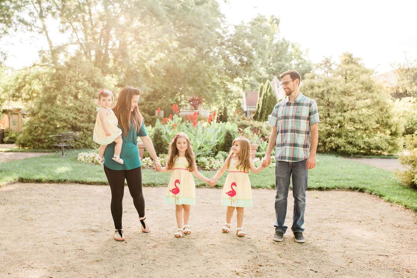 A couple stands while holding their daughters' hands in a garden.