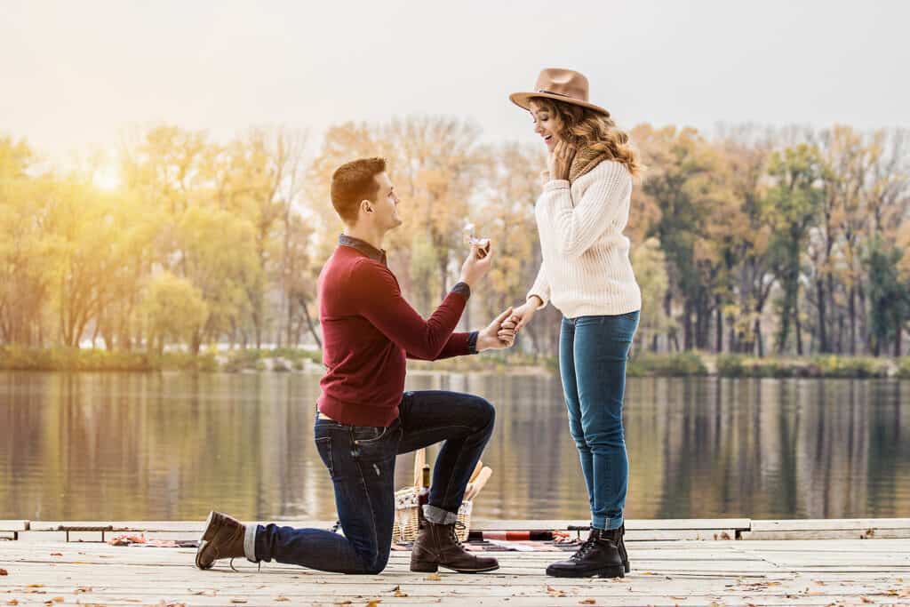 A man is on his knee proposing to a woman by a lake.