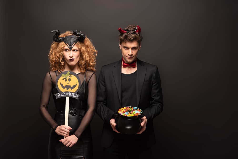 A couple in Halloween costumes poses against a black wall in the background.