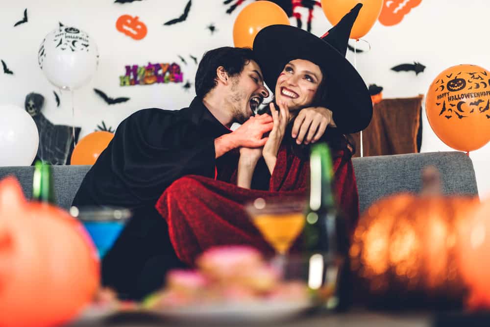 A couple is sitting while dressed in Halloween costumes surrounded by orange balloons.