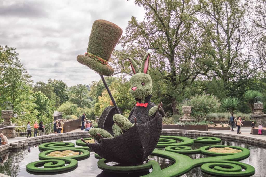 Green statue made out of plants of a bunny with a hat sitting in an umbrella.