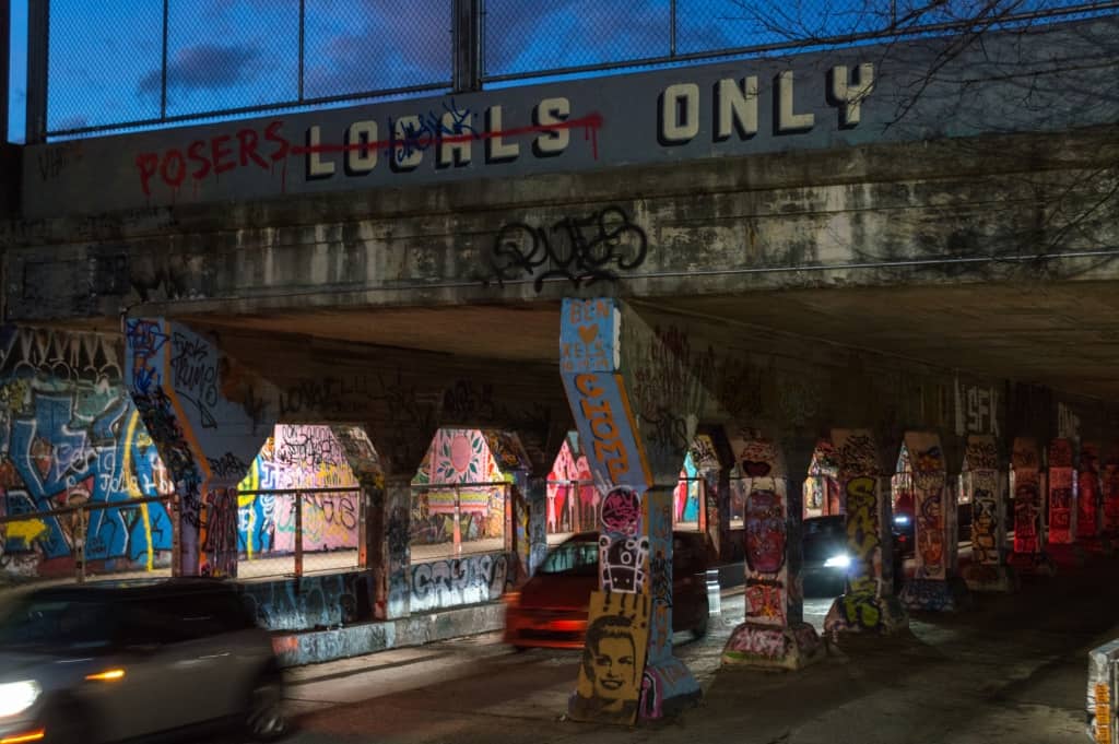 Cars driving under a bridge covered in graffiti.