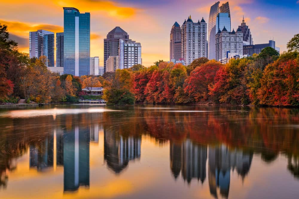 A city skyline under an orange and purple sky in fall is reflected on a serene lake.