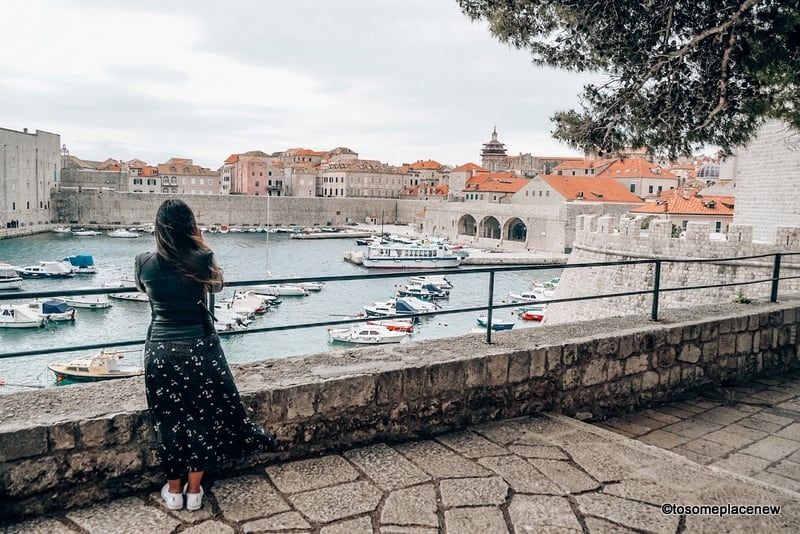 A woman stands looking out at boats floating in the harbor under a grey sky.