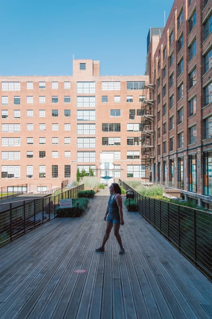 A woman stands on an empty walkway surrounded by brick buildings.