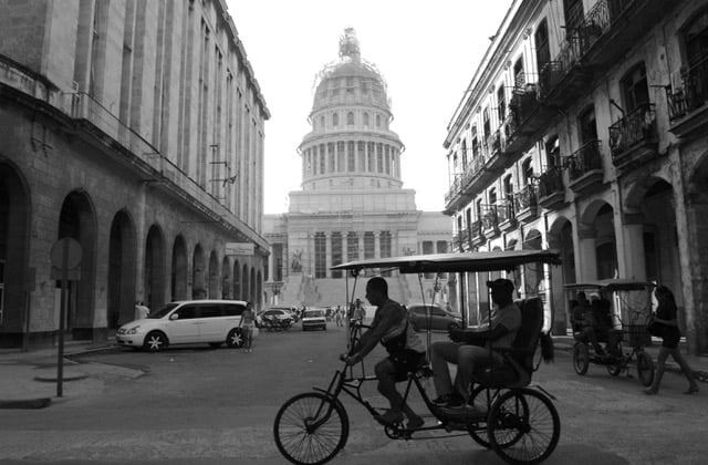 Two people on a bicycle drive by a big government building.
