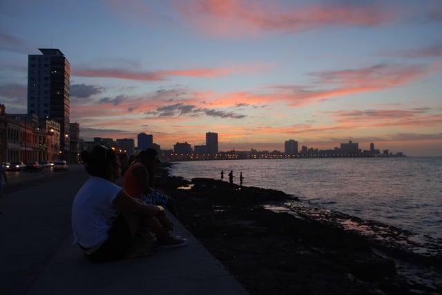 People sit and watch the sunset over the water under a blue and pink sky.