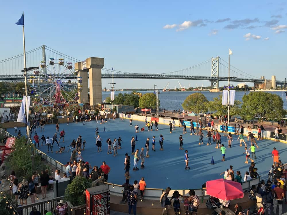 People roller skate outdoors next to the water. A bridge crosses the water in the background.