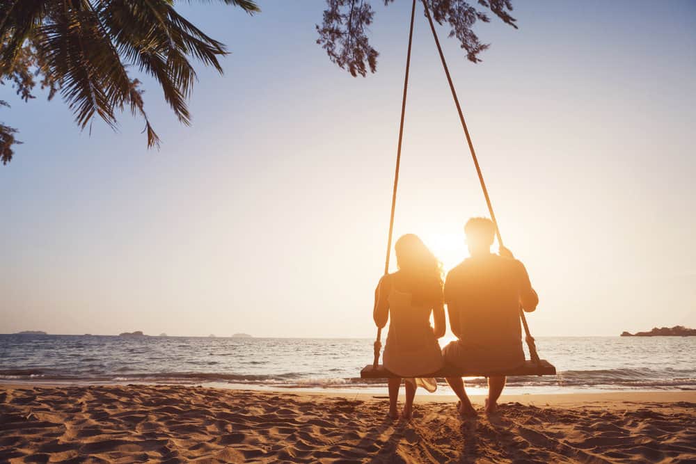 A couple swings on a beach at sunset with palm trees above them.