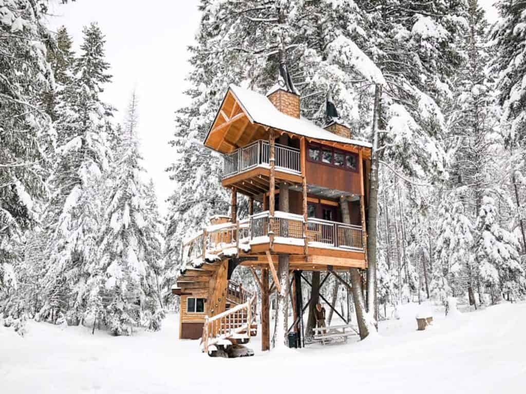 A tall cabin in the woods is surrounded by snow.