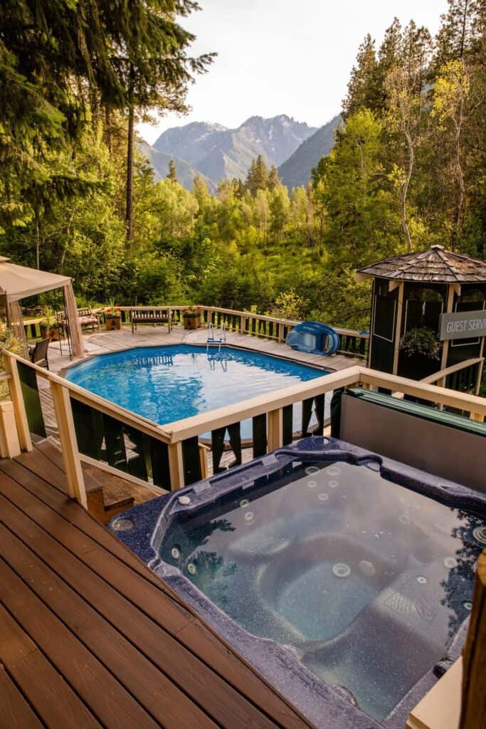 A hot tub is outdoors with a pool behind it. In the distance are mountains and trees.