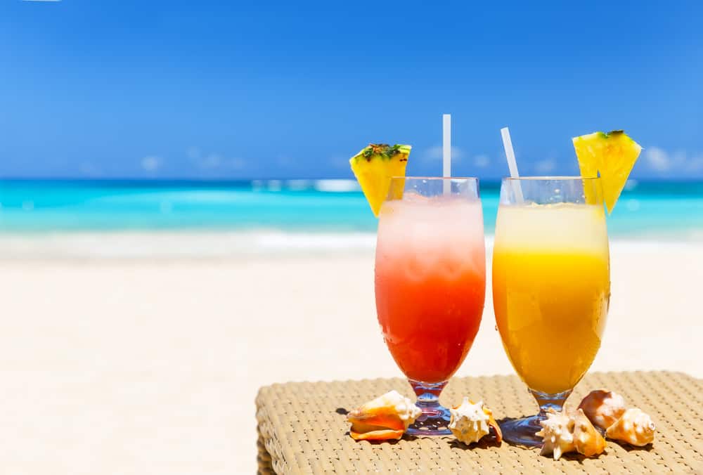 A pink and yellow drink with a pineapple wedge sits on a table with a blurred out ocean behind.