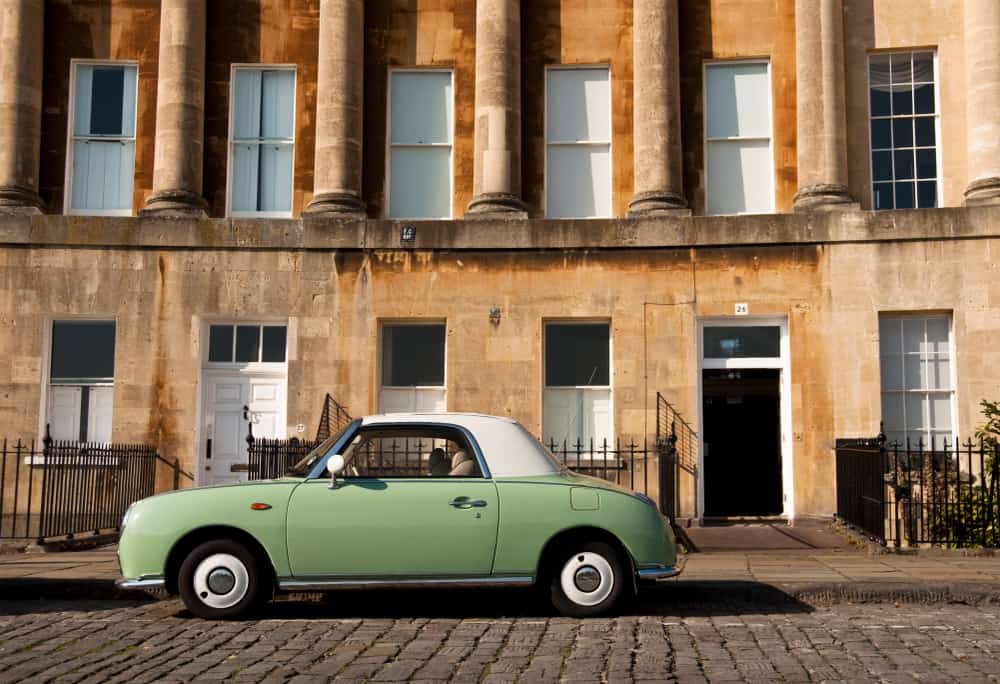 An old-school green car is parked on a cobblestone street by a tan-colored stone building.
