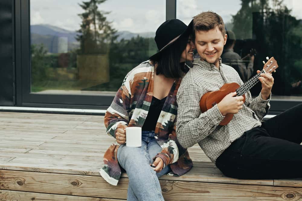 A couple sits on a porch laughing. The woman drinks coffee and is nuzzling her face against the man who is playing the ukelele.