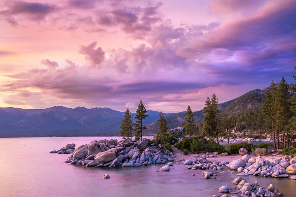 A calm lake is surrounded by mountains under a purple sky.