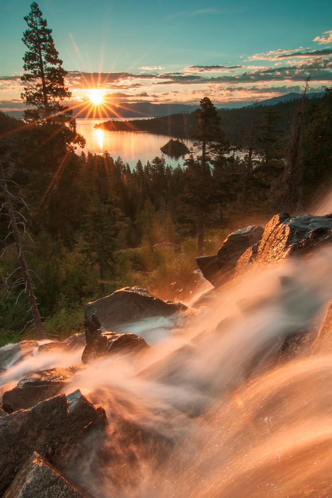 Water rushes down a rocky mountainside. A lake is seen in the distance where the sun is shining through.