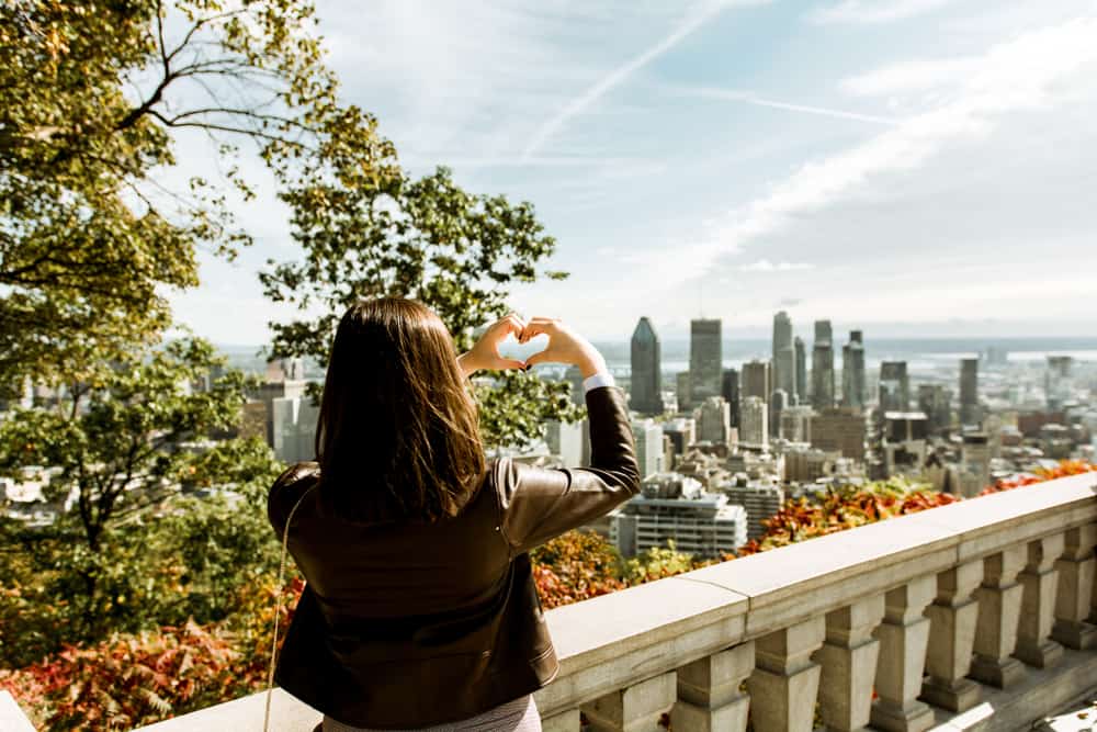 A woman makes a heart shape with her hands while looking out at a view of the city skyline.