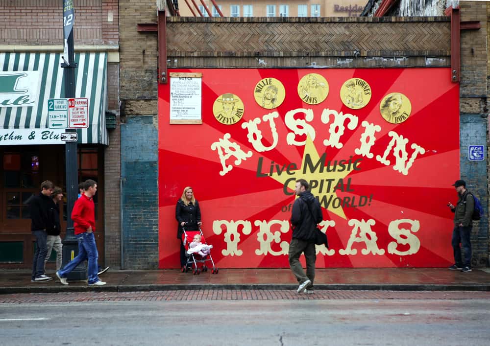 People walk by a mural that\'s bright red that says Austin Texas, Live music capital of the world.