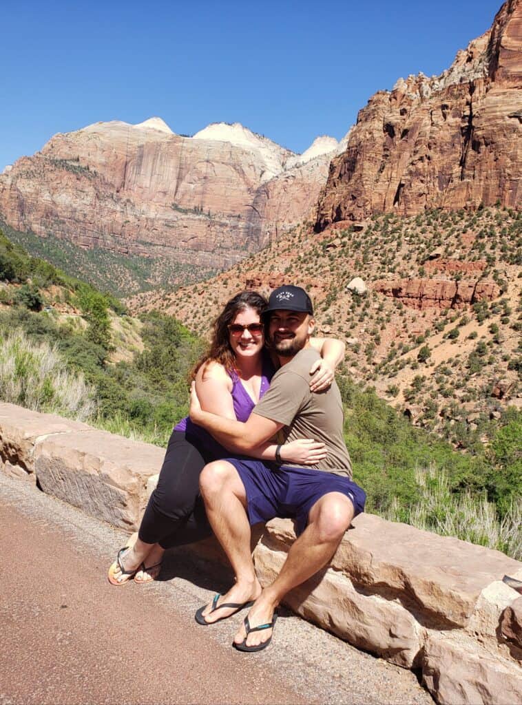 A couple sits on a perch by the round with tall rocky mountains behind them.