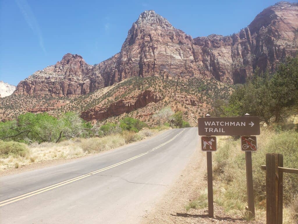 An empty paved road leading through an area surrounded by tall, rocky mountains. In the foreground is a sign that says \"Watchman Trail\" with an arrow pointing right.