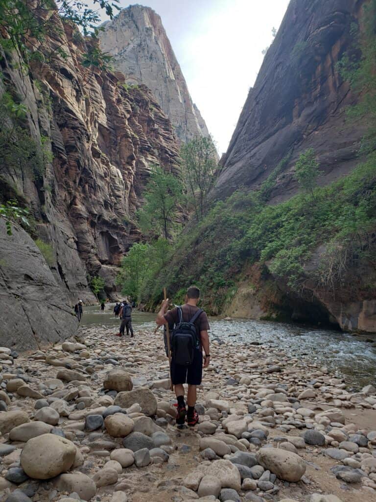 Hikers walking along the rocky shore of a canyon floor with a small river to the right. 
