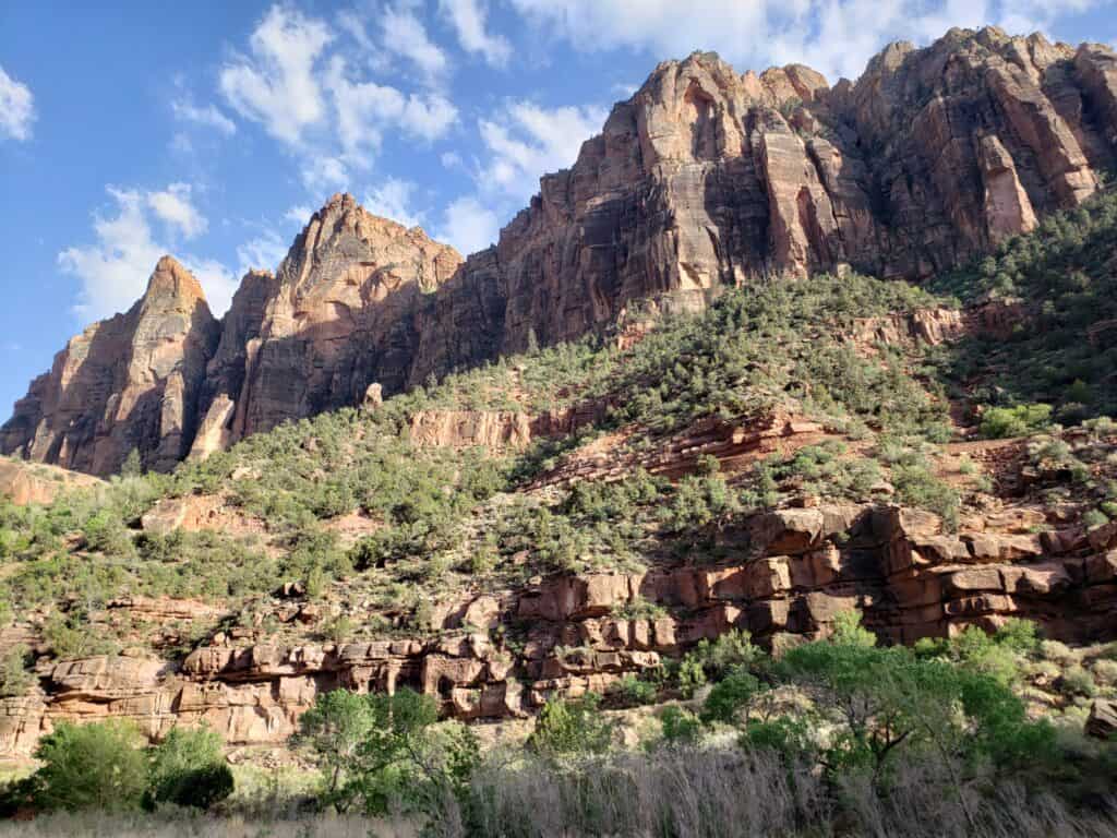 View from the bottom of a canyon looking up at a large rock formation with grass and trees along the side. 