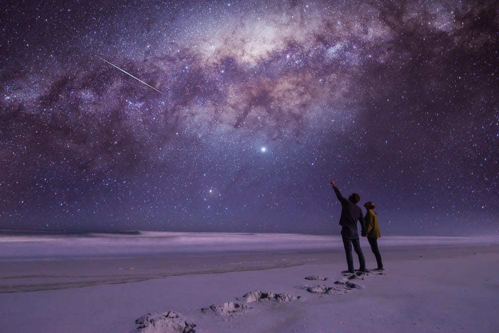 A couple stargazes in a beautiful open spot in the same, with a blue and purple milky way above them.