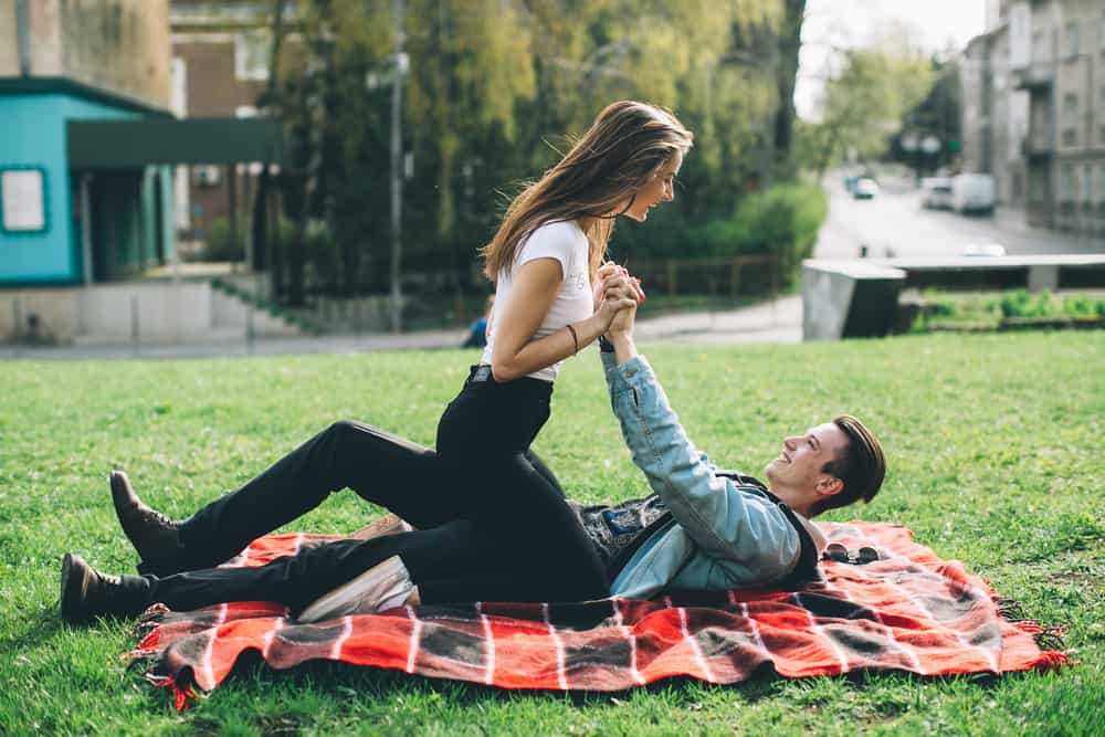 A woman sits on top of a man on a red and black plaid blanket in a field.