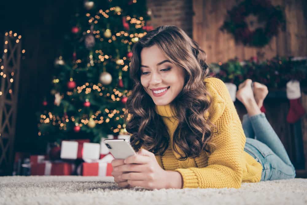 Woman texting in front of Christmas tree and smiling reading flirty Christmas texts