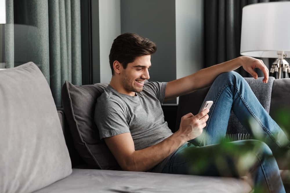 A man smiles on the couch while reading a text.